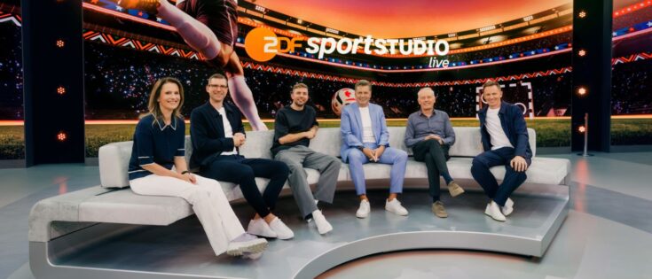 Sechs Personen sitzen auf einem Sofa im ZDF Sportstudio, mit einem Fußballstadion-Hintergrund und WM-Atmosphäre.