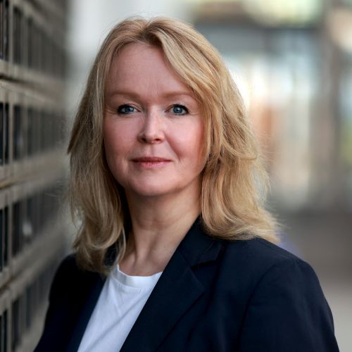 Porträt einer Frau mit blonden Haaren, die in einem blauen Blazer lächelt, vor einem unscharfen Hintergrund.