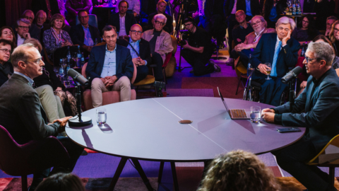 Zwei Männer sitzen an einem runden Tisch und führen ein Gespräch vor einem Publikum in einem gut beleuchteten Raum.