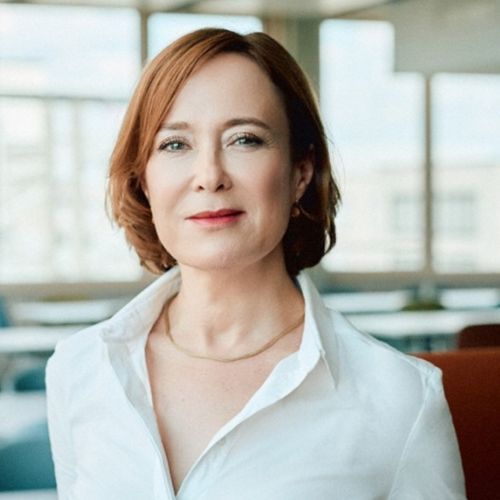 Porträt von Tatjana Dreyer in einem hellen Raum, mit kurzem, rotem Haar und weißem Hemd, lächelnd in die Kamera blickend.