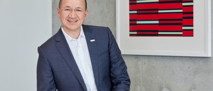 Portrait von Prof. Dr. Christof Ehrhart in Anzug, lächelnd vor einer modernen Wand mit abstrakter Kunst.