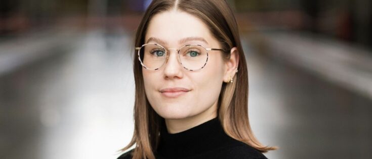 Porträt einer jungen Frau mit Brille, braunen Haaren und schwarzem Pullover, lächelnd in einem modernen Innenraum.