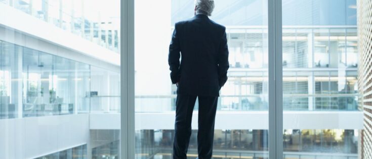 Geschäftsmann in Anzug steht vor einer Glaswand in einem modernen Bürogebäude und blickt nach draußen.