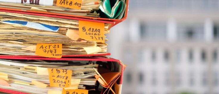 Ein Stapel unordentlicher Aktenordner im Vordergrund, unscharfes Bürogebäude im Hintergrund.