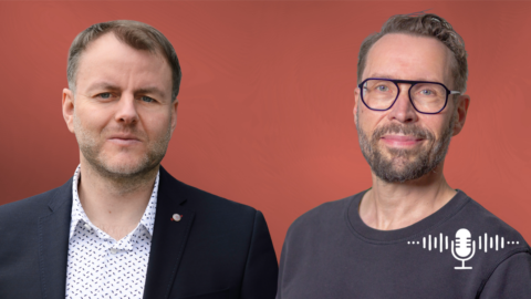 Zwei Männer stehen vor einem rötlichen Hintergrund, einer in einem Anzug, der andere in einem T-Shirt, beide lächeln.