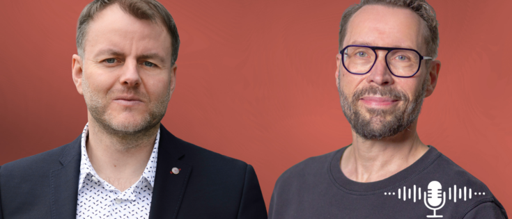 Zwei Männer stehen vor einem rötlichen Hintergrund, einer in einem Anzug, der andere in einem T-Shirt, beide lächeln.