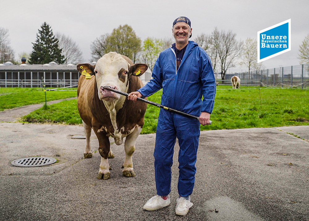 Mann in blauer Arbeitskleidung steht lächelnd neben einer Kuh auf einer Wiese, im Hintergrund Bäume und ein Stall.