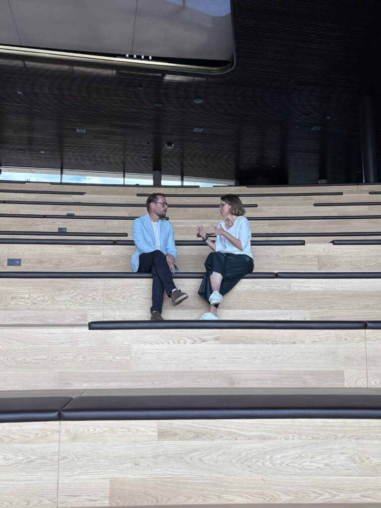Zwei Personen sitzen auf Holzstufen in einem modernen Gebäude und führen ein Gespräch.