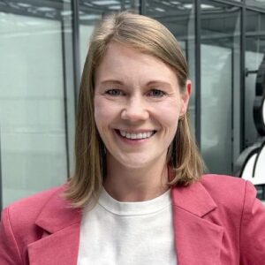Porträt einer lächelnden Frau mit blonden Haaren, die einen rosa Blazer trägt, vor einer modernen Glasarchitektur.