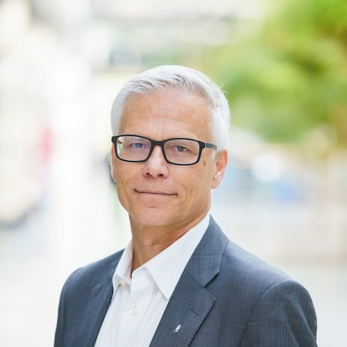 Mann mit grauen Haaren und Brille, trägt einen Anzug, steht lächelnd im Freien mit unscharfem Hintergrund.