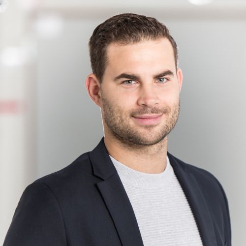 Porträt eines Mannes mit kurzem, dunklem Haar, in einem schwarzen Blazer und grauem Pullover, lächelnd vor einem hellen Hintergrund.