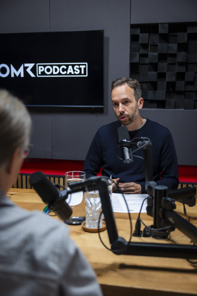Zwei Personen sitzen an einem Tisch in einem Podcast-Studio, Mikrofone sind aufgestellt, eine Person spricht.