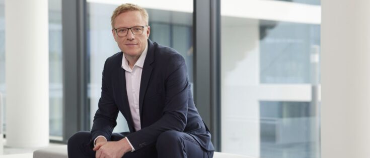 Mann in Anzug sitzt auf einer Bank vor großen Fenstern in modernem Gebäude.