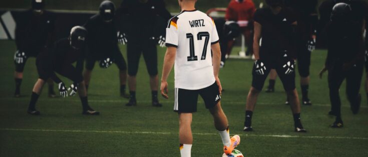 Foto aus einer Adidas-Kampagne: Florian Wirtz steht mit dem Rücken zur Kamera, sein rechter Fuß ruht auf einem Fußball. Vor sich steht eine Reihe schwarz gekleideter Spieler mit Handschuhen und schwarzen Helmen.