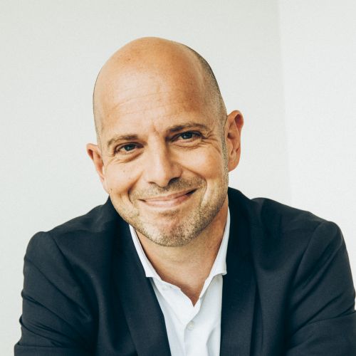 Porträt eines lächelnden Mannes mit Glatze, in einem dunklen Anzug und weißem Hemd, vor hellem Hintergrund.