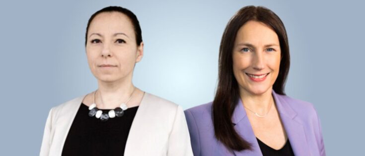 Zwei Frauen stehen nebeneinander vor einem hellblauen Hintergrund, eine in weißem Blazer, die andere in lila Jacke.