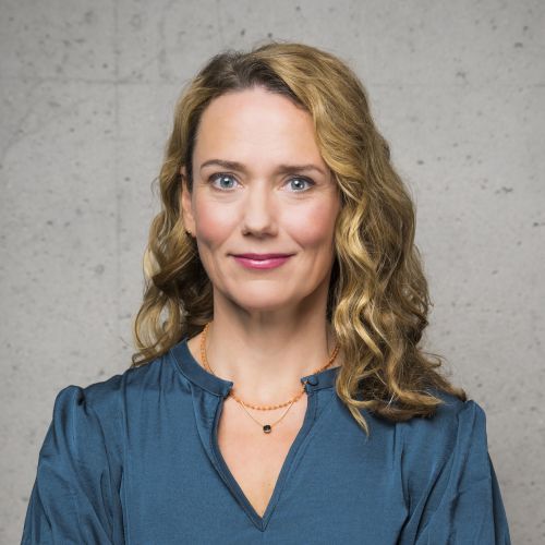 Porträt einer Frau mit lockigem, blondem Haar, die in einem blauen Oberteil vor einer grauen Wand steht.