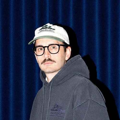 Mann mit Brille und Schnurrbart trägt einen grauen Hoodie und eine weiße Cap vor einem blauen Hintergrund.