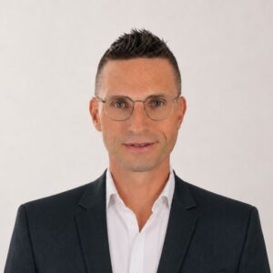 Mann mit Brille und Anzug, lächelt vor neutralem Hintergrund.