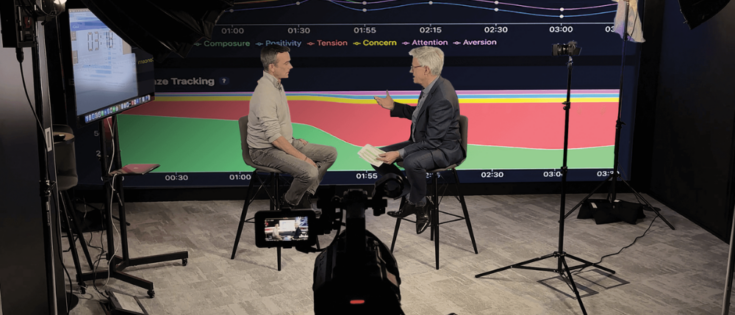Zwei Männer sitzen in einem Studio, diskutieren vor einem grafischen Hintergrund mit Linien und Zeitmarkierungen.