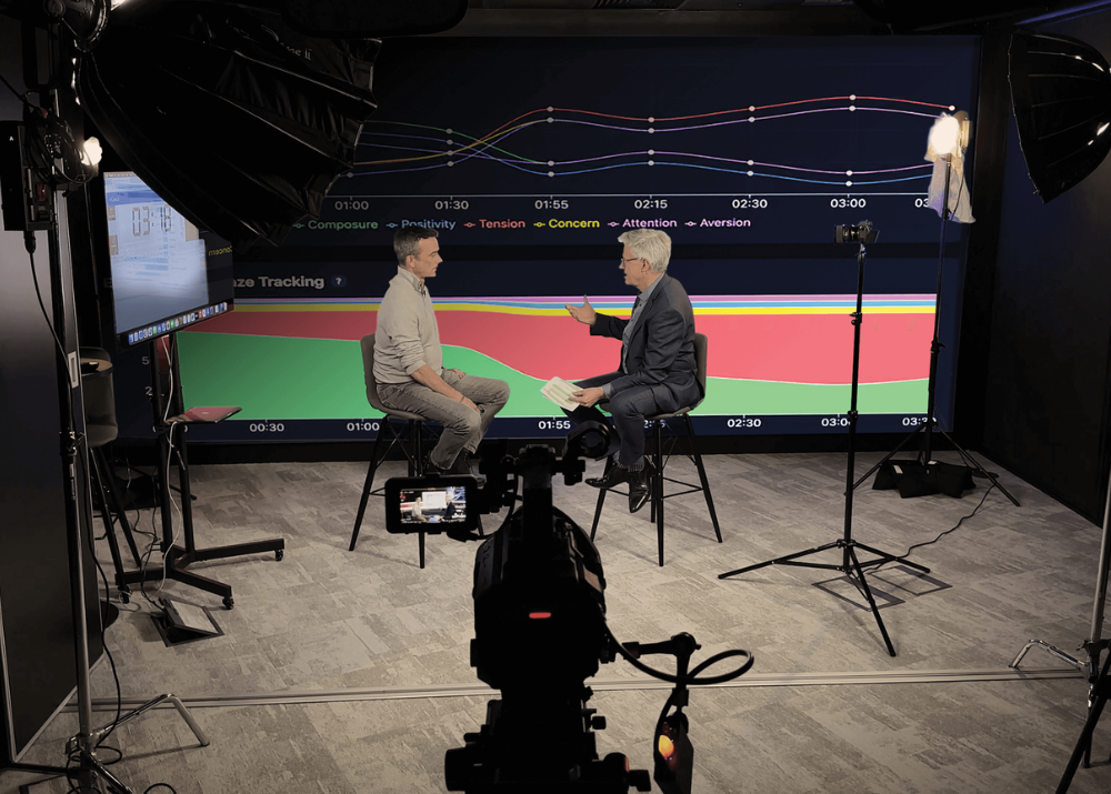 Zwei Männer sitzen in einem Studio, diskutieren vor einem grafischen Hintergrund mit Linien und Zeitmarkierungen.