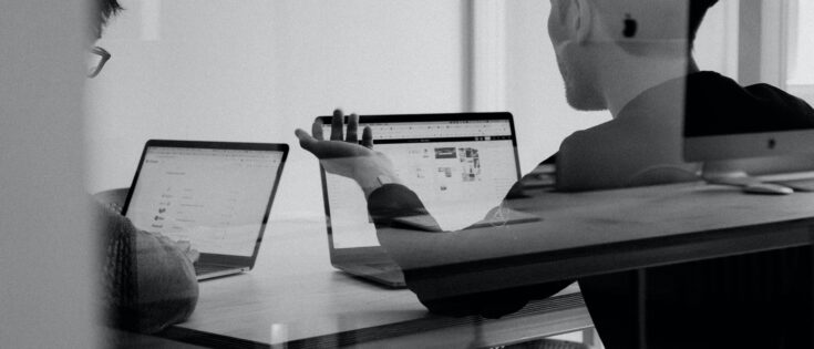 Zwei Personen sitzen an einem Tisch mit Laptops und diskutieren, der Raum ist hell und modern gestaltet.