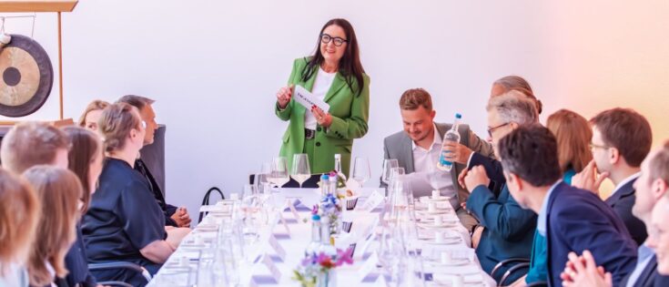 Frau in grüner Jacke spricht vor einer Gruppe von Menschen an einem langen Tisch, umgeben von Gläsern und Blumen.