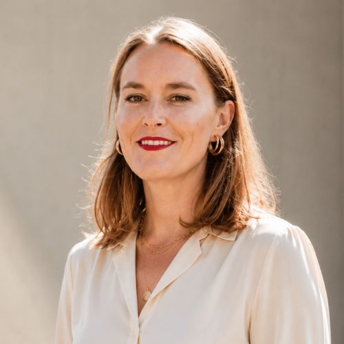 Porträt einer Frau mit schulterlangen, dunkelblonden Haaren und weißer Bluse vor einem hellen Hintergrund.