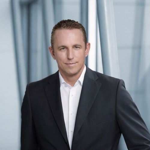 Porträt eines Mannes in einem Anzug mit kurzem, blondem Haar, vor einem modernen, hellen Hintergrund.