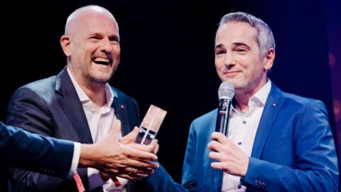 Johannes Winter und Oliver Claas von Leica Camera nehmen den Preis entgegen. © Anika Nowak Zwei Männer in Anzügen lächeln, während einer einen Preis überreicht. Festliche Atmosphäre auf einer Bühne.