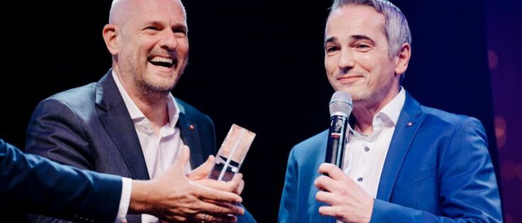 Johannes Winter und Oliver Claas von Leica Camera nehmen den Preis entgegen. © Anika Nowak Zwei Männer in Anzügen lächeln, während einer einen Preis überreicht. Festliche Atmosphäre auf einer Bühne.