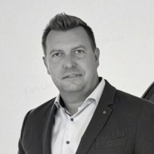 Mann in Anzug mit kurzem, blondem Haar, lächelt in die Kamera vor einem neutralen Hintergrund.