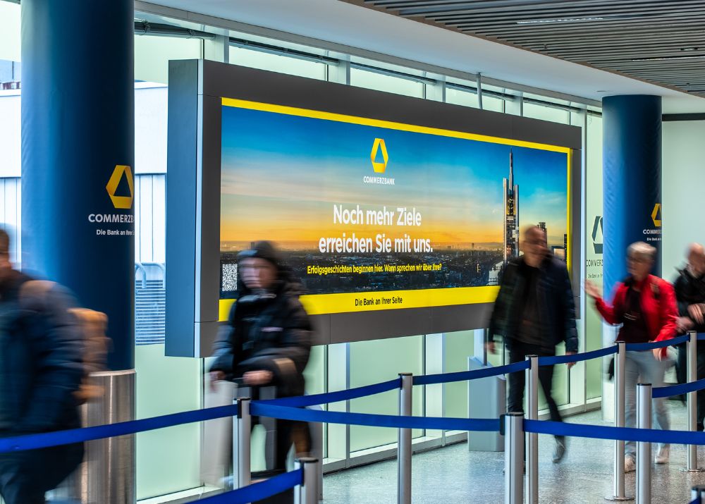 Großes Werbebild der Commerzbank mit Skyline und Text über Ziele am Flughafen, umgeben von wartenden Menschen.