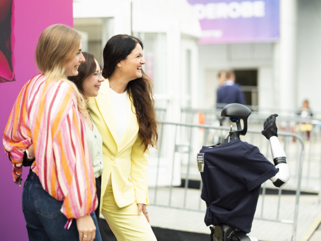 © Mirella Frangella Drei Frauen posieren lächelnd für ein Foto mit einem Roboter, der ein T-Shirt trägt, vor einem bunten Hintergrund.