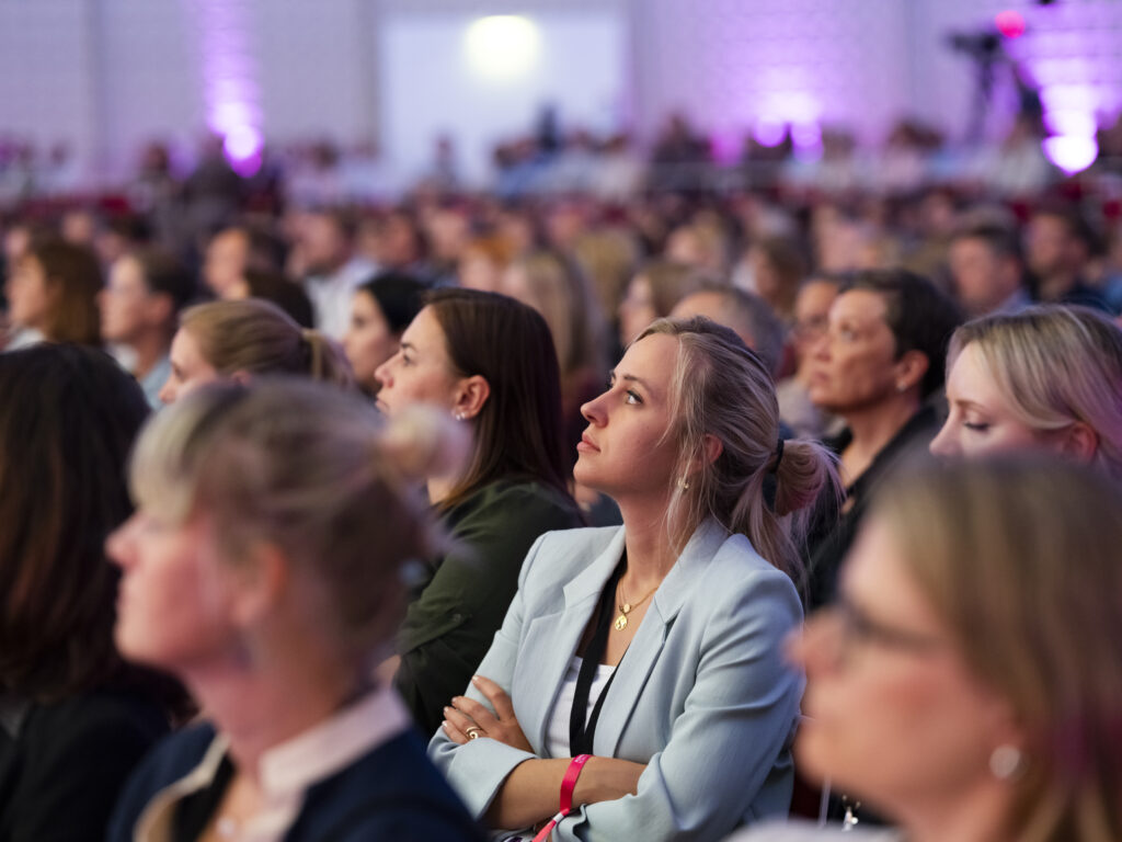 © Mirella Frangella Audience gathered at an indoor event; foreground shows a woman in a light blue blazer with arms crossed, attentively watching.