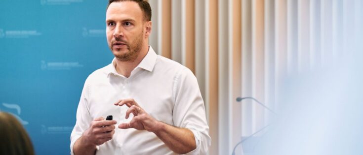 Mann in weißem Hemd spricht mit einem Mikrofon, im Hintergrund eine Präsentation und beige Wände.