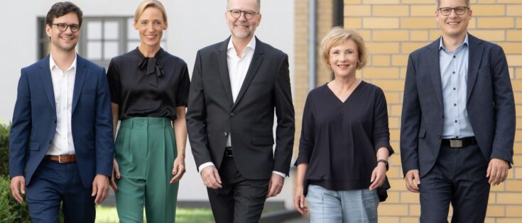 Fünf Personen in formeller Kleidung gehen lächelnd auf einem Gehweg entlang, umgeben von moderner Architektur.