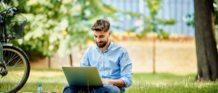 Junger Mann sitzt im Park auf dem Gras und arbeitet an einem Laptop, neben ihm steht ein Fahrrad.