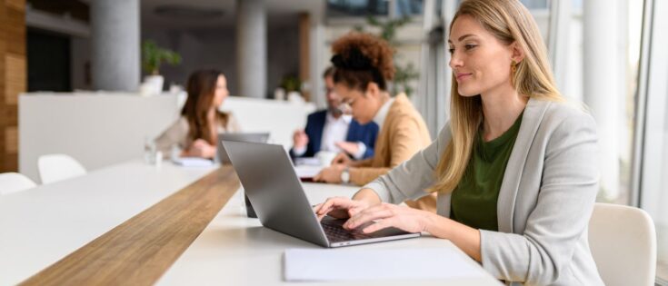 Frau analysiert Bericht am Laptop in einem modernen Besprechungsraum, umgeben von Kollegen.