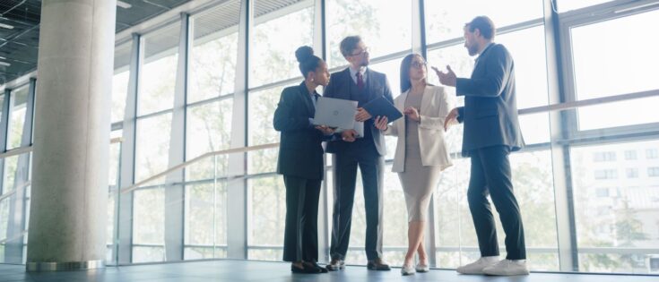 Business-Team in einem hellen Büroflur diskutiert Projektupdates und überprüft digitale Berichte auf Laptop und Tablet.