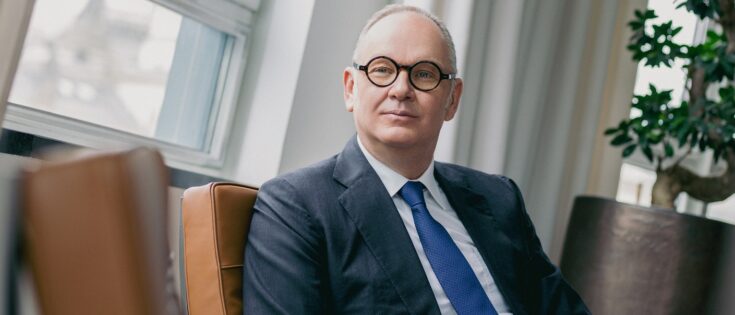 Mann mit Brille und Anzug sitzt entspannt in einem modernen Raum mit Fenster und Pflanzen im Hintergrund.