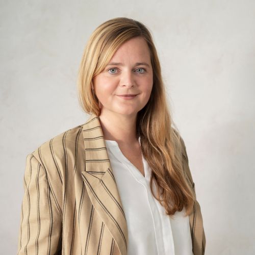 Porträt einer Frau mit langen, blonden Haaren, die einen gestreiften Blazer und ein weißes Hemd trägt, vor einem neutralen Hintergrund.