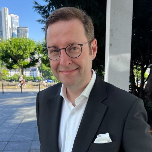 Mann mit Brille und Anzug, lächelt vor urbaner Kulisse mit Bäumen und Gebäuden im Hintergrund.