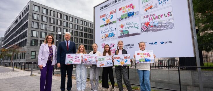 Kinder halten Zeichnungen vor einem großen Plakat, das auf Rücksichtnahme im Straßenverkehr hinweist.
