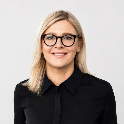 Porträt einer lächelnden Frau mit blonden Haaren und Brille, in schwarzem Hemd vor neutralem Hintergrund.