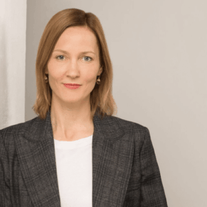 Frau mit blonden Haaren in einem grauen Blazer, steht vor einem neutralen Hintergrund und schaut direkt in die Kamera.