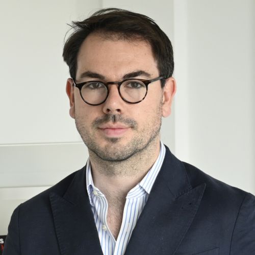 Porträt eines Mannes mit Brille, dunklem Anzug und gestreifter Hemdkragen, vor neutralem Hintergrund.