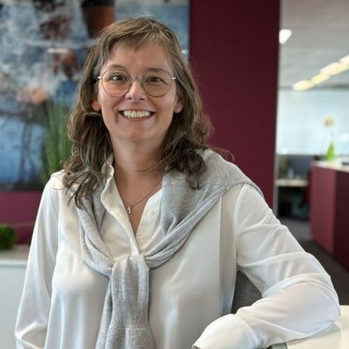 Frau mit Brille und grauem Pullover über der Schulter lächelt in einem modernen Büro.