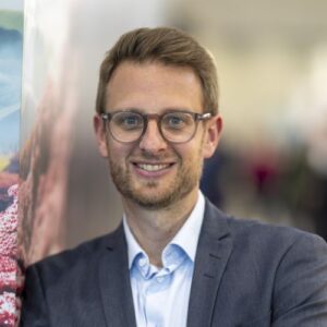 Mann mit Brille und Anzug lächelt vor einem unscharfen Hintergrund.