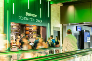 Jägermeister-Promotionsstand am Flughafen mit Kunden und Regalen voller Flaschen, grüne Beleuchtung im Hintergrund.